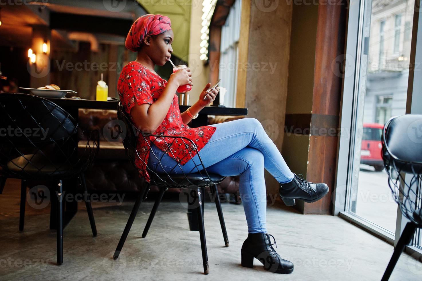 Stylish african woman in red shirt and hat posed indoor cafe and drinking strawberry lemonade with phone at hand. photo