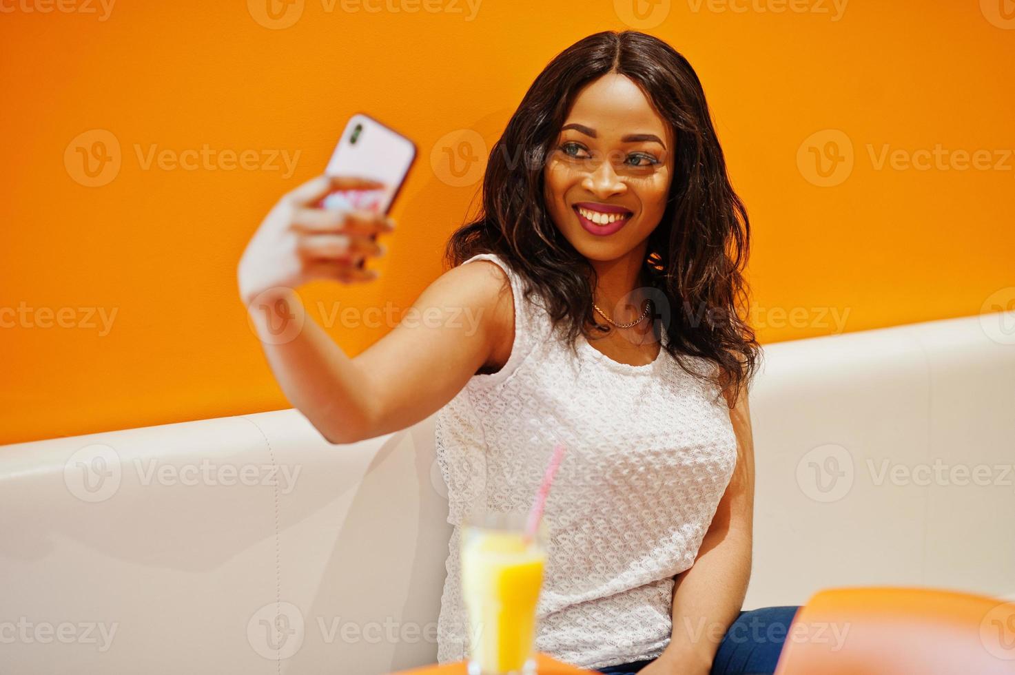 African woman sitting on cafe against orange wall with pineapple juice and mobile phone in hands, she making selfie. photo