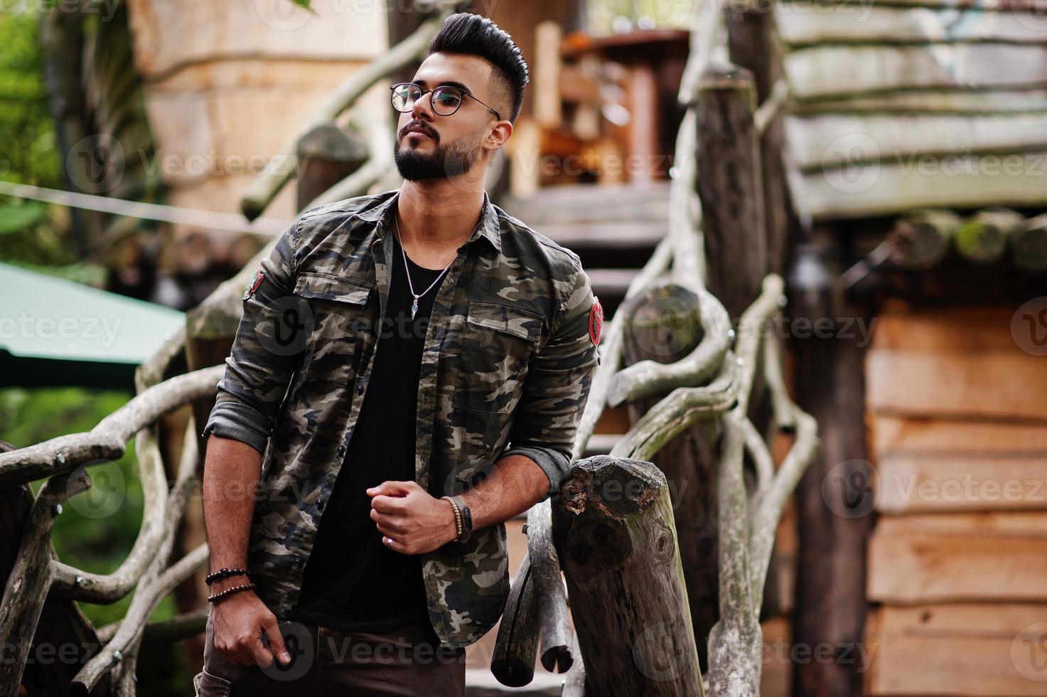 Awesome beautiful tall ararbian beard macho man in glasses and military jacket posed outdoor against wooden stairs. photo