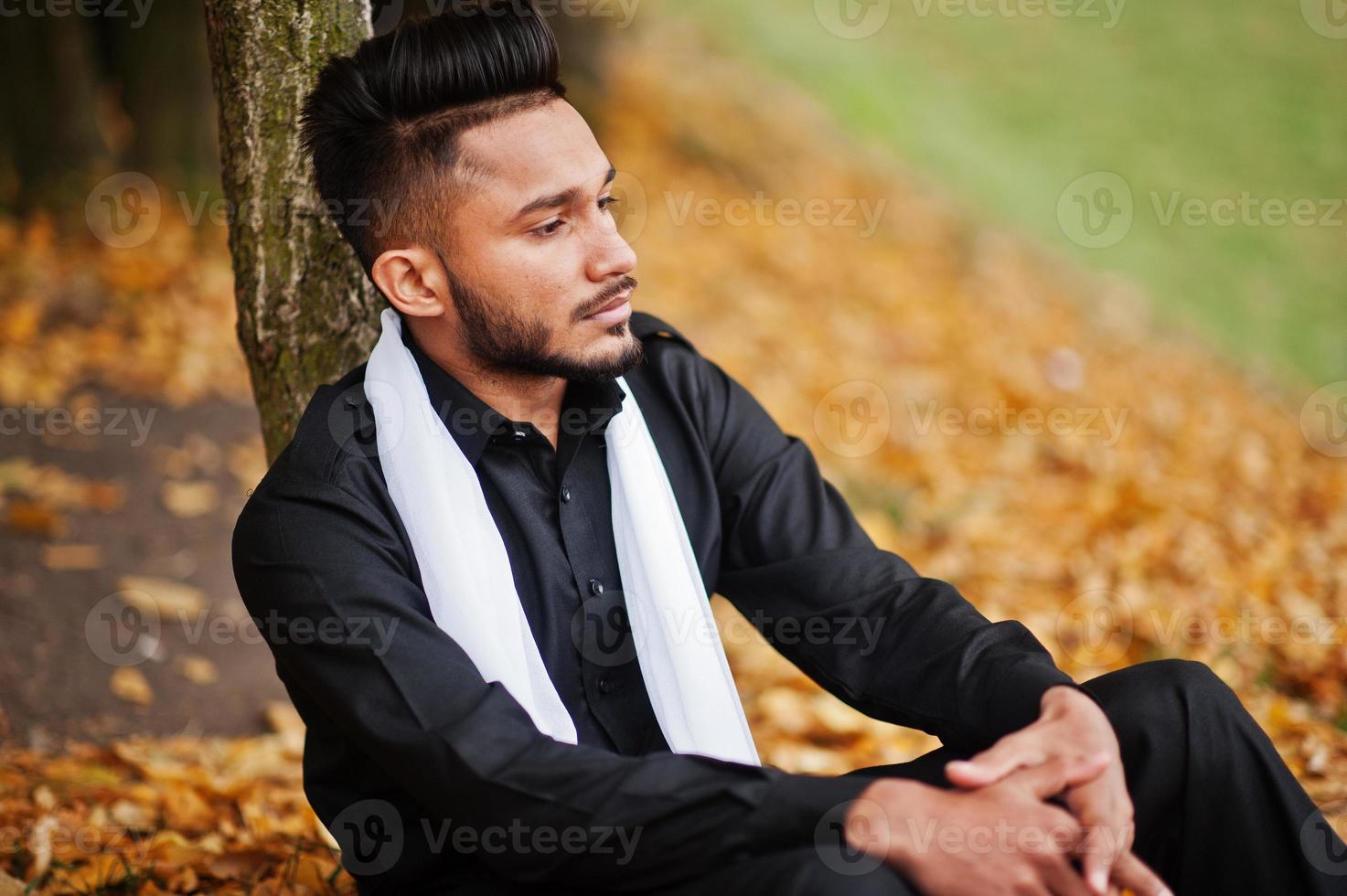 Indian stylish man in black traditional clothes with white scarf posed outdoor against yellow autumn leaves tree. photo