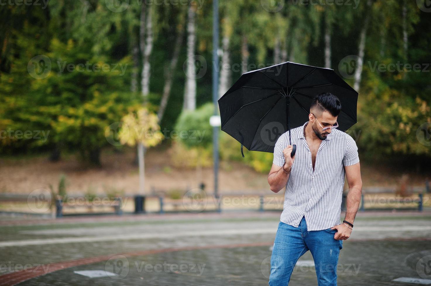 Fashionable tall arab beard man wear on shirt, jeans and sunglasses with umbrella posed at rain on park square. photo