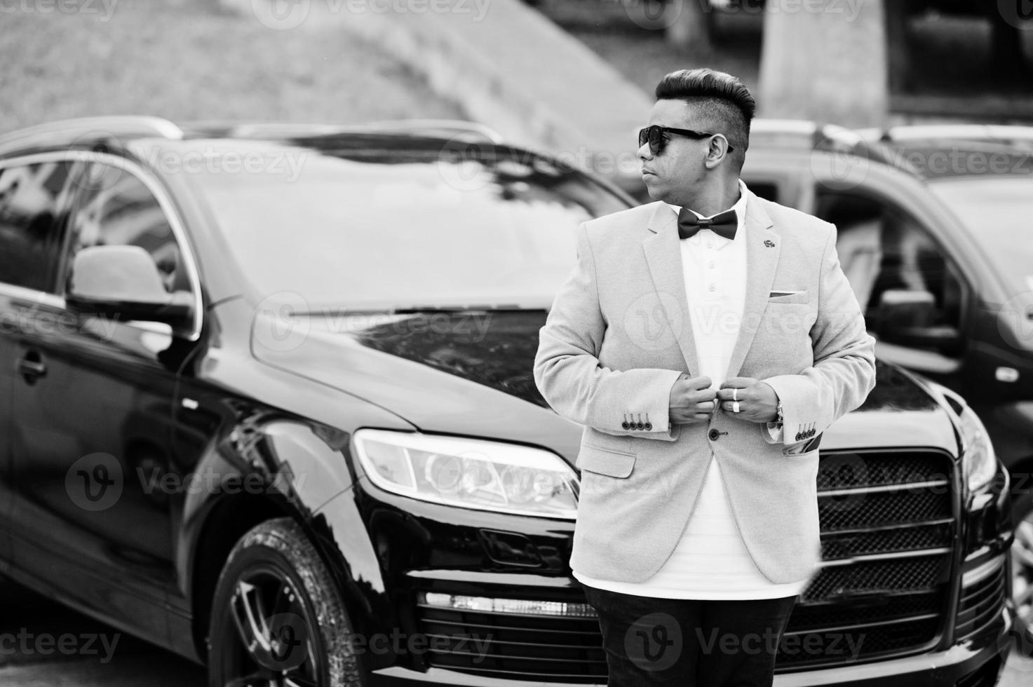 Stylish arabian man in jacket, bow tie and sunglasses against black suv car. Arab rich businessman. photo