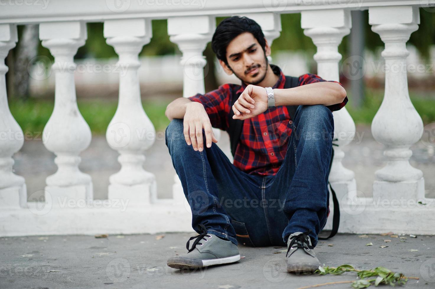 Young indian student man at checkered shirt and jeans with backpack posed on evening city against column and looking at his watches. photo