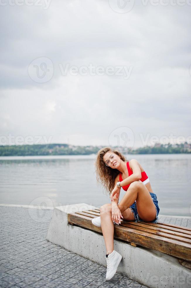 chica sexy modelo rizada en top rojo, pantalones cortos de mezclilla, anteojos y zapatillas posadas en un banco contra el lago. foto