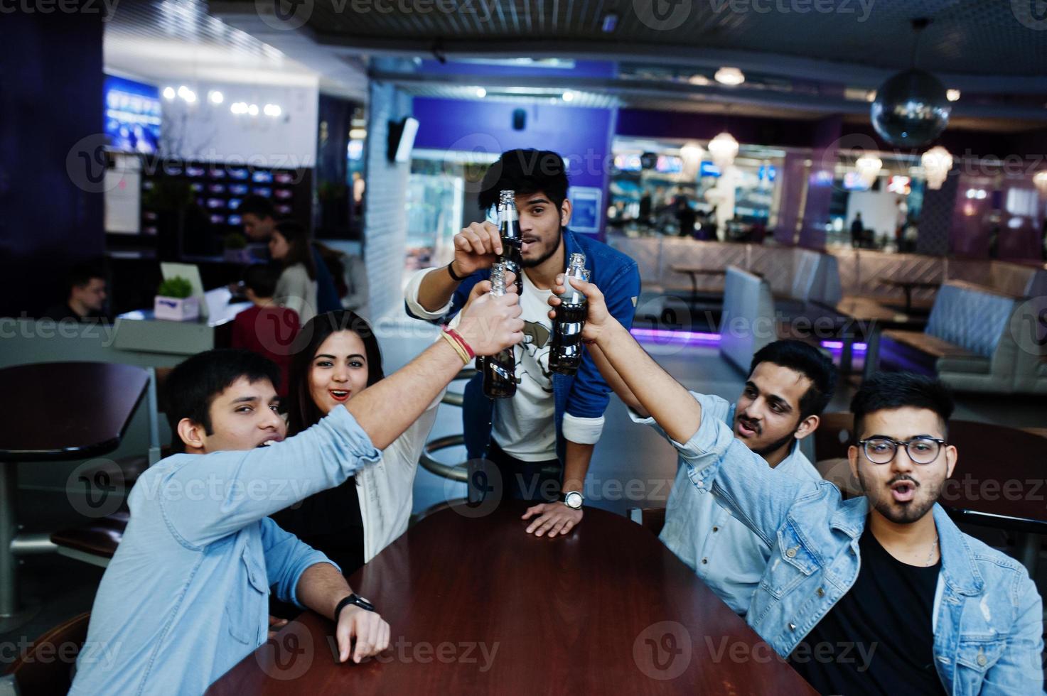 Group of stylish asian friends wear on jeans sitting at table and cheering with soda bottles in club. photo