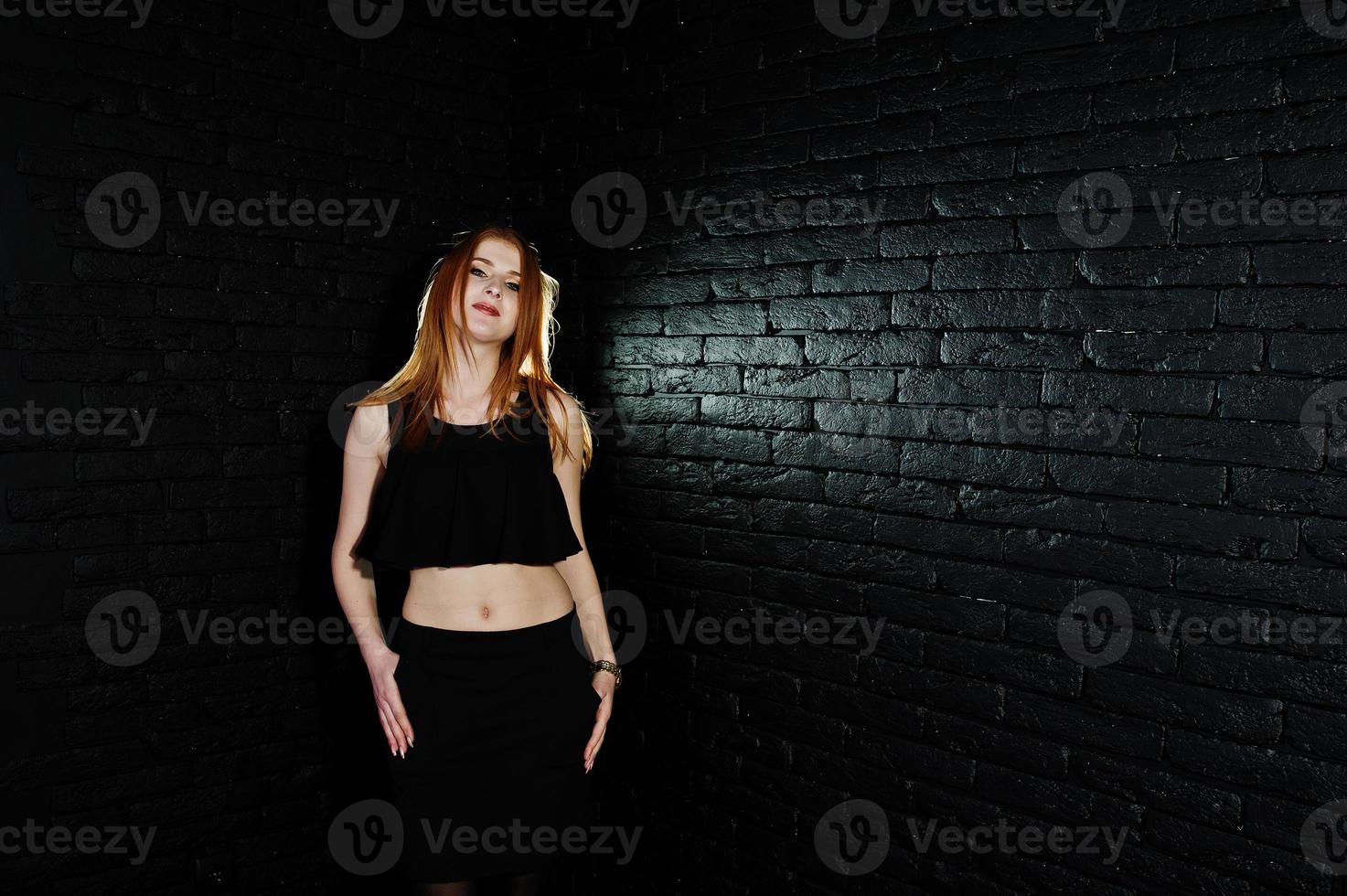 Portrait of a beautiful redheaded girl in black top and black skirt posing in the studio next to the brick wall. photo