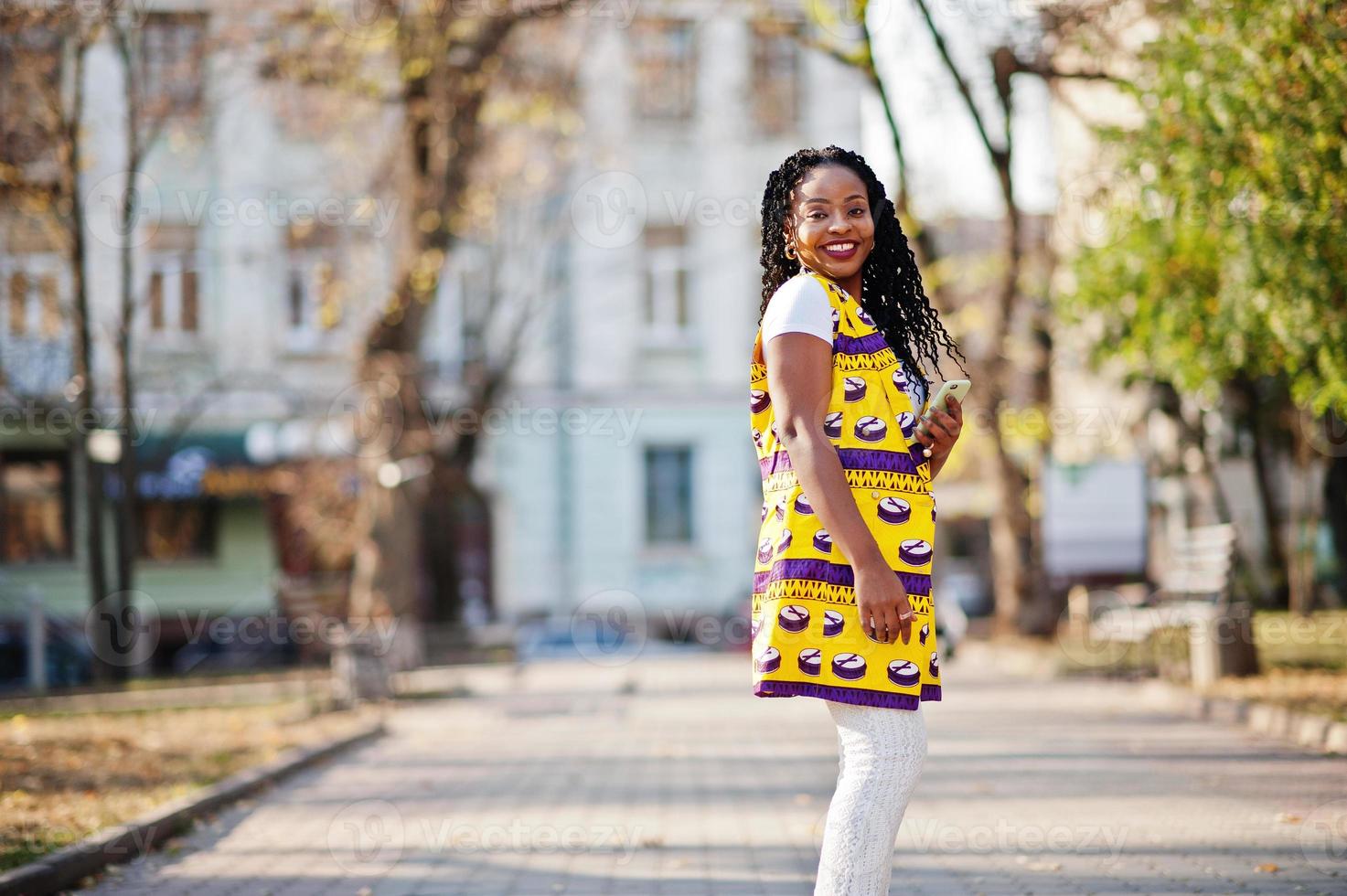 Stylish african american women in yellow jacket posed on street at sunny day with mobile phone at hand. photo