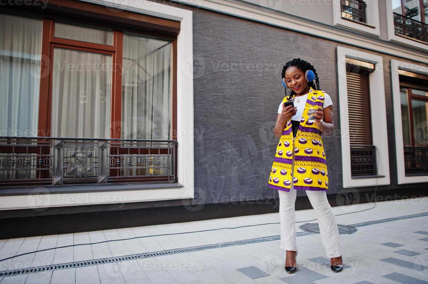 Stylish african american women in yellow jacket posed and earphones on street against modern building with hot drink in disposable paper cup. photo