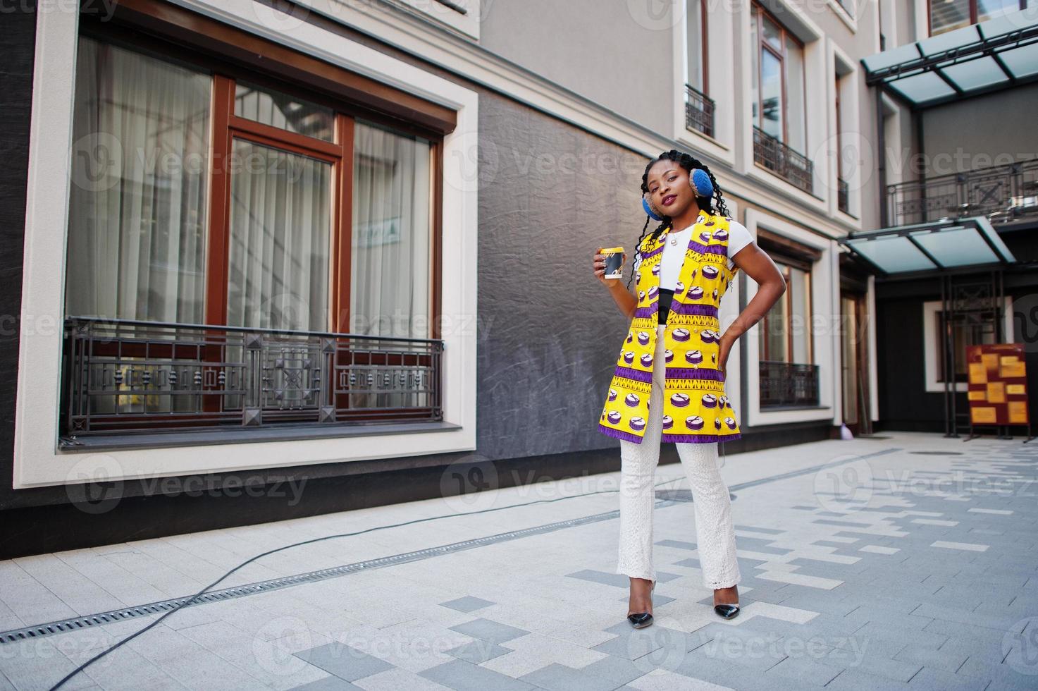 Stylish african american women in yellow jacket posed and earphones on street against modern building with hot drink in disposable paper cup. photo
