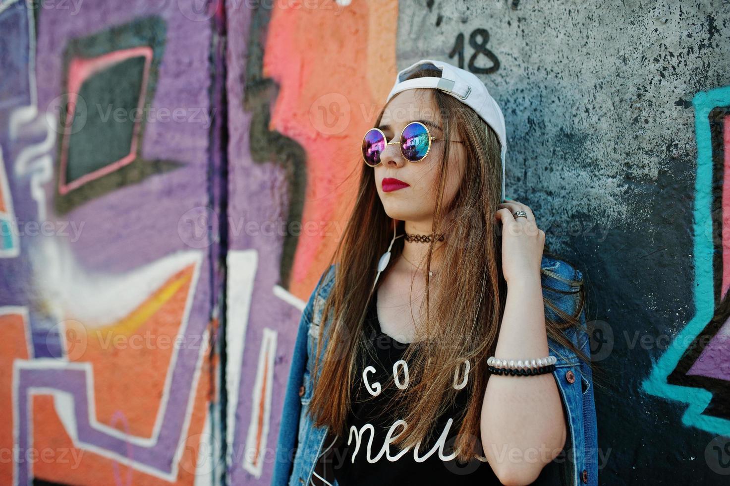 elegante chica hipster casual con gorra, gafas de sol y jeans, escuchando música desde auriculares de teléfono móvil contra una gran pared de graffiti. foto
