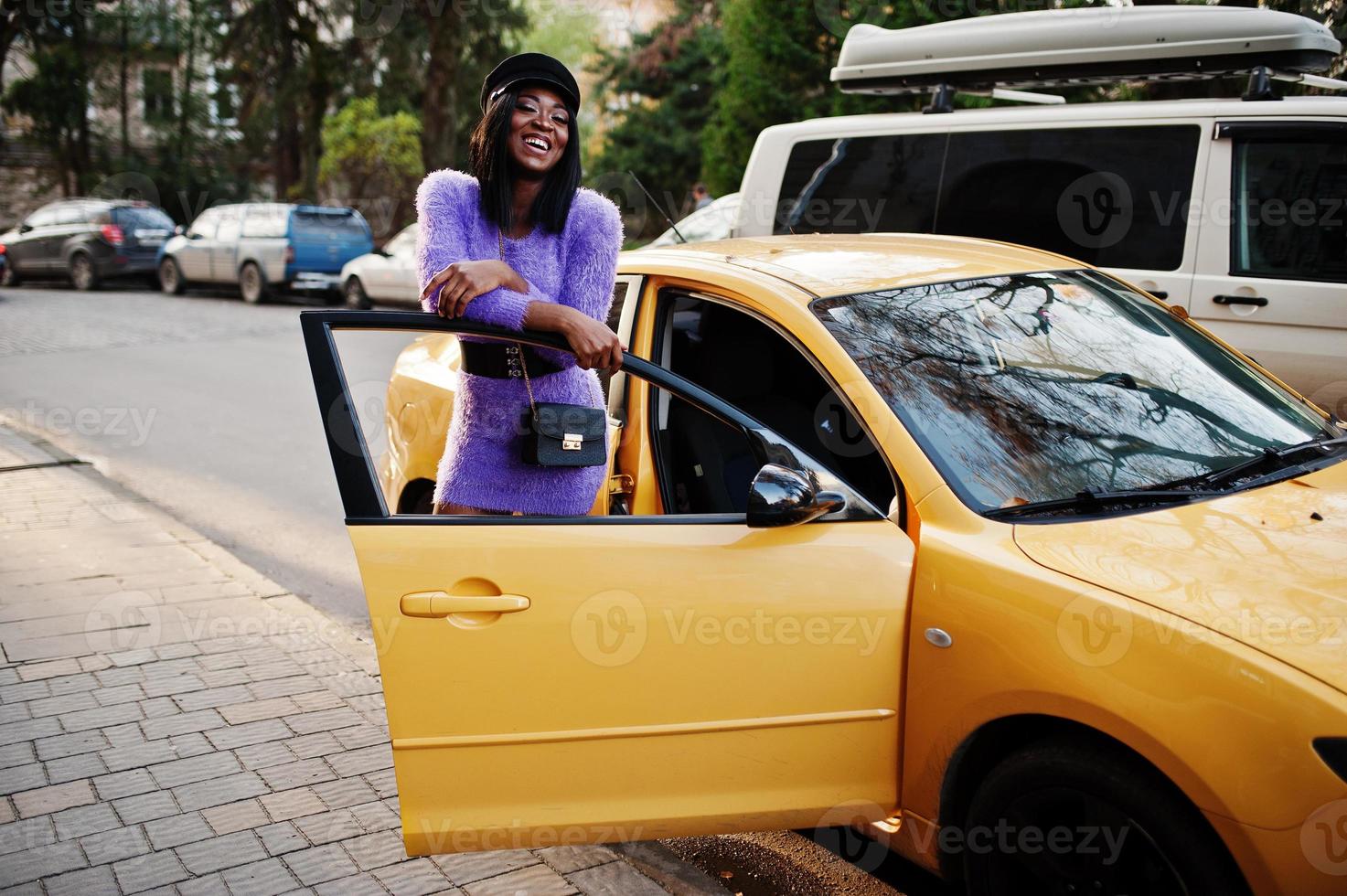 African american woman at violet dress and cap posed at yellow car. photo