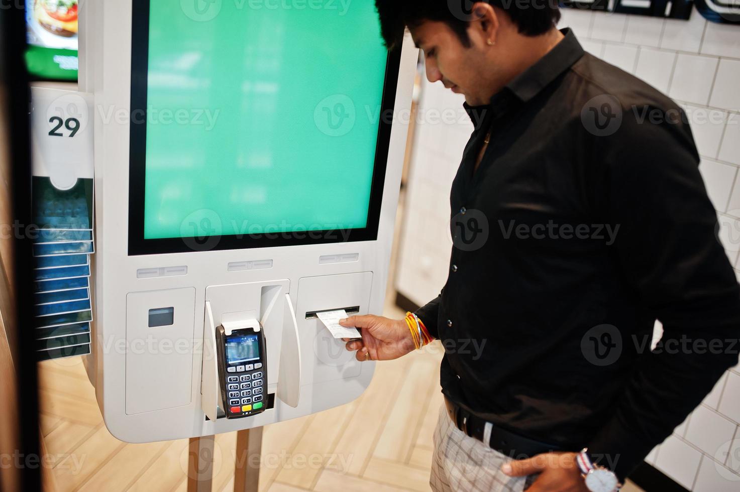 Indian man customer at store place orders and pay through self pay floor kiosk for fast food, payment terminal. His taking fiscal check. photo