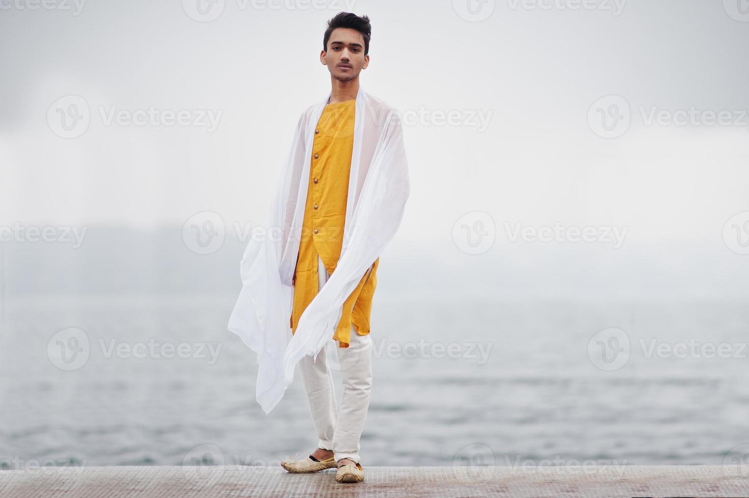 Indian stylish man in yellow traditional clothes with white scarf posed outdoor against foggy pier of the sea. photo