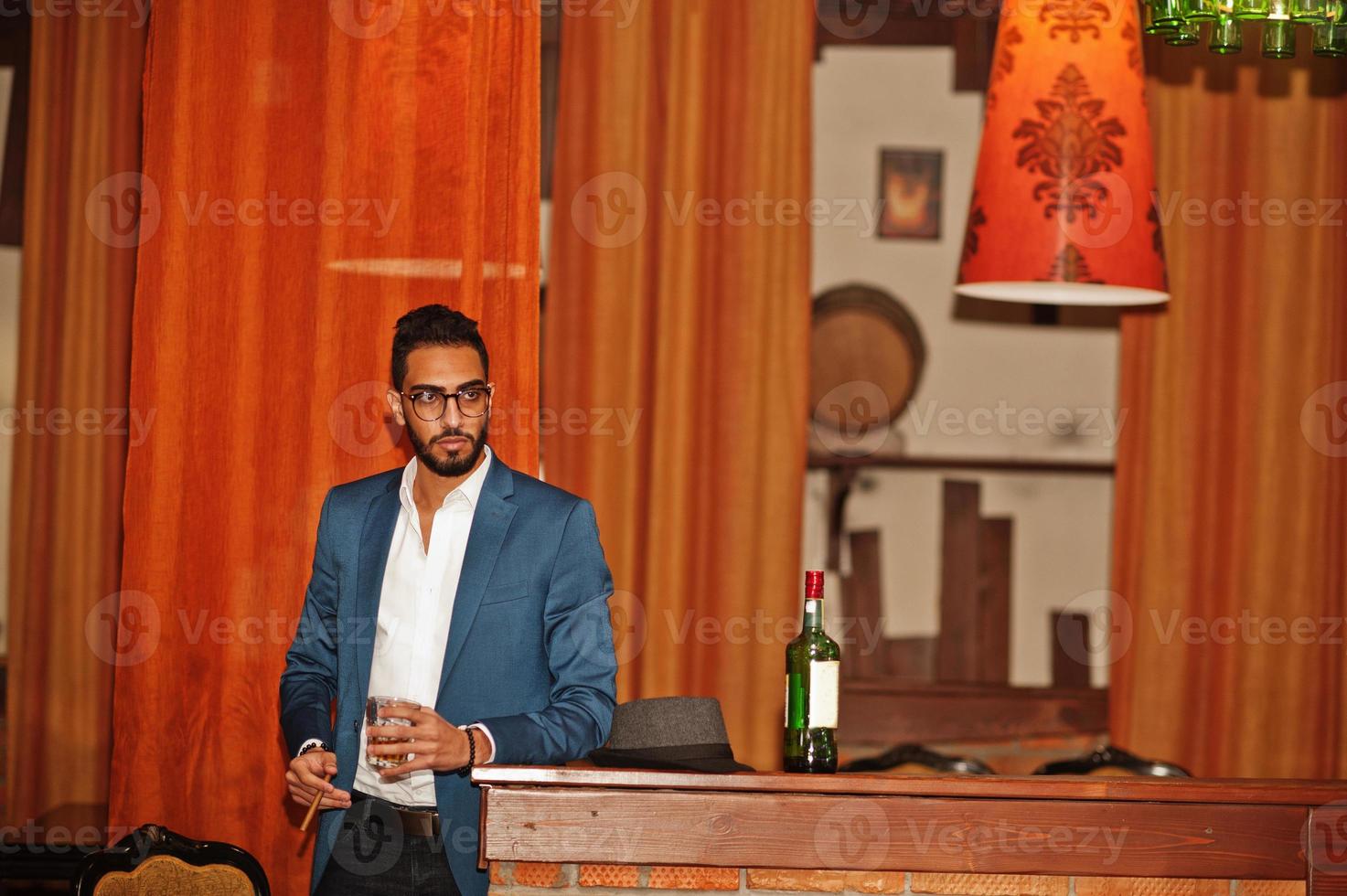 Handsome well-dressed arabian man with glass of whiskey and cigar posed at pub. photo