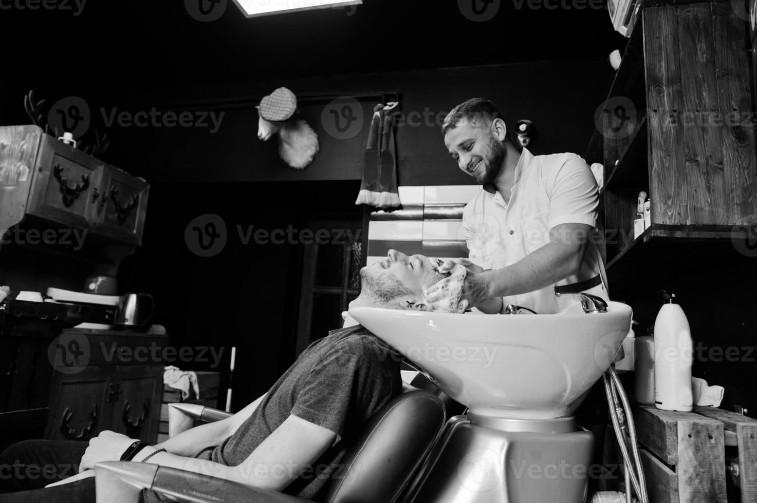 Young bearded man washing head by hairdresser while sitting in chair at barbershop. Barber soul. photo