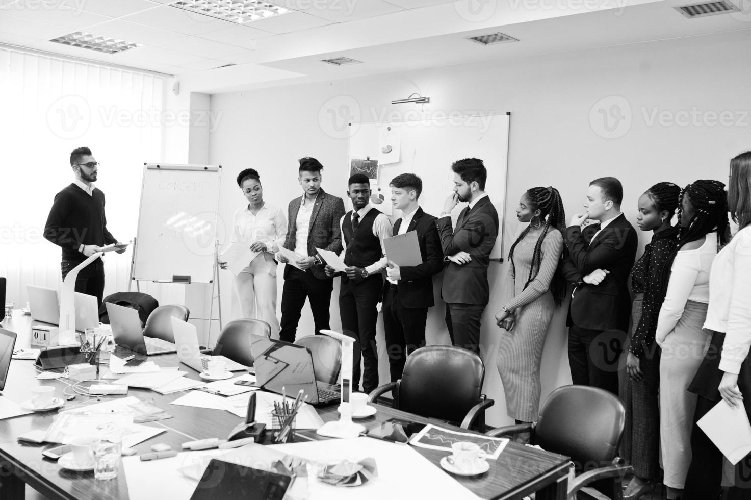 Mixed race business coach presenting report standing near whiteboard pointing on sales statistic shown on diagram and chart teach diverse company members gathered together in conference room. photo