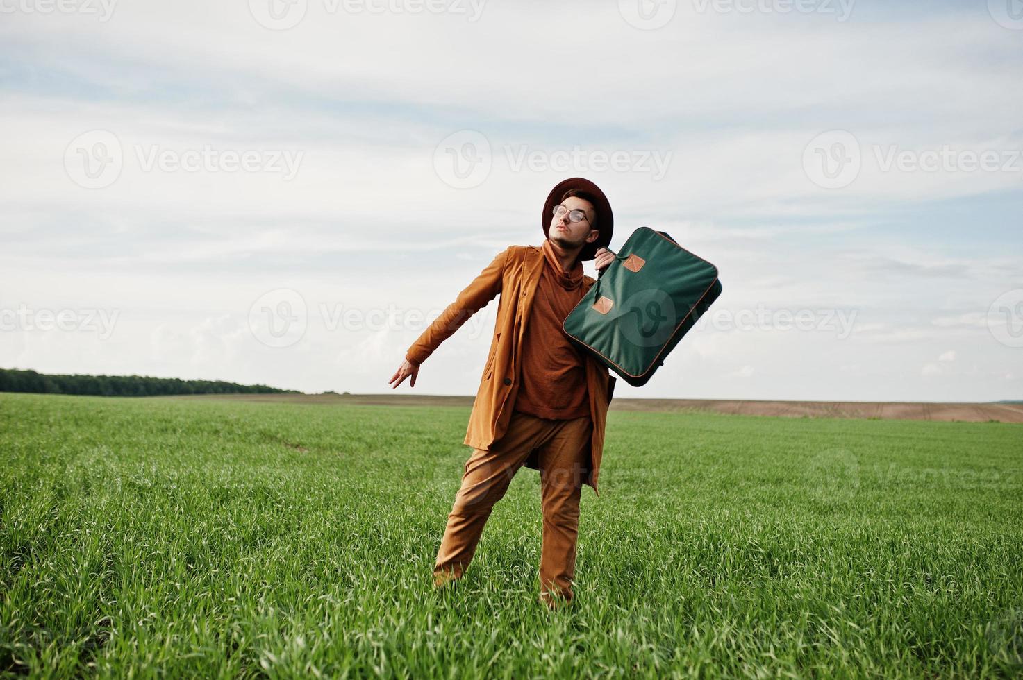 Stylish man in glasses, brown jacket and hat with bag posed on green field. photo