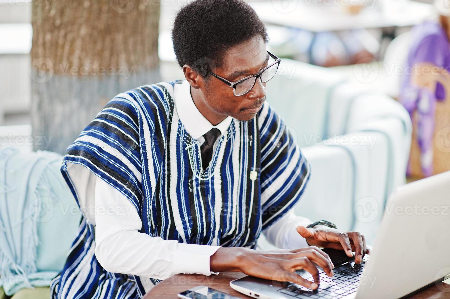 African man in traditional clothes and glasses sitting behind laptop at outdoor caffe and working. photo