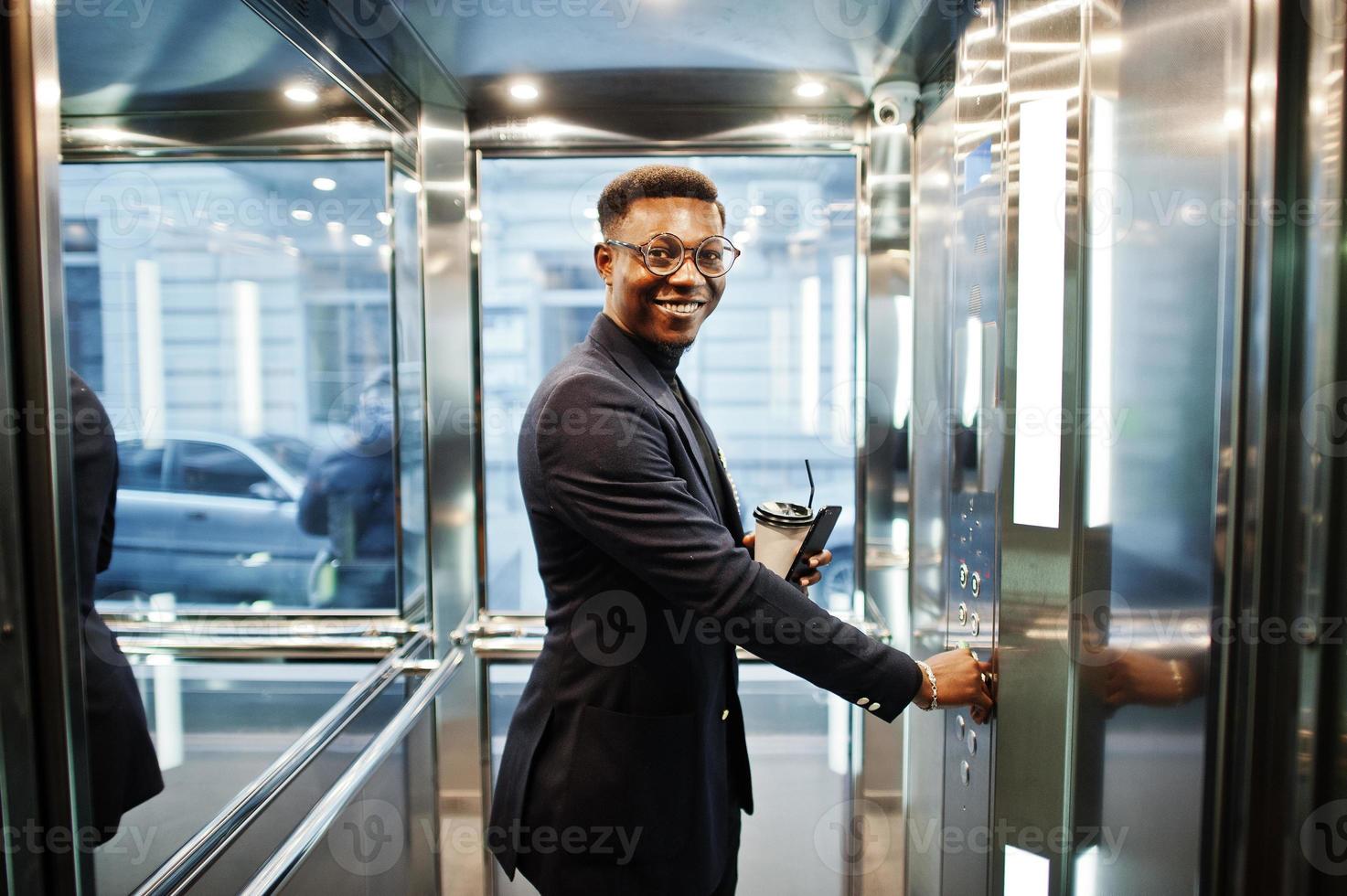 hombre afroamericano de moda con traje y gafas con teléfono móvil y una taza de café en las manos posado dentro del ascensor. foto
