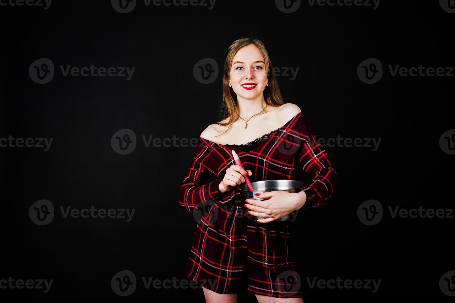 Young housewife in checkered dress with saucepan and kitchen spoon isolated on black background. photo