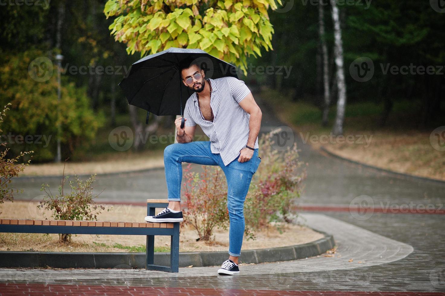 Fashionable tall arab beard man wear on shirt, jeans and sunglasses with umbrella posed at rain on park square. photo