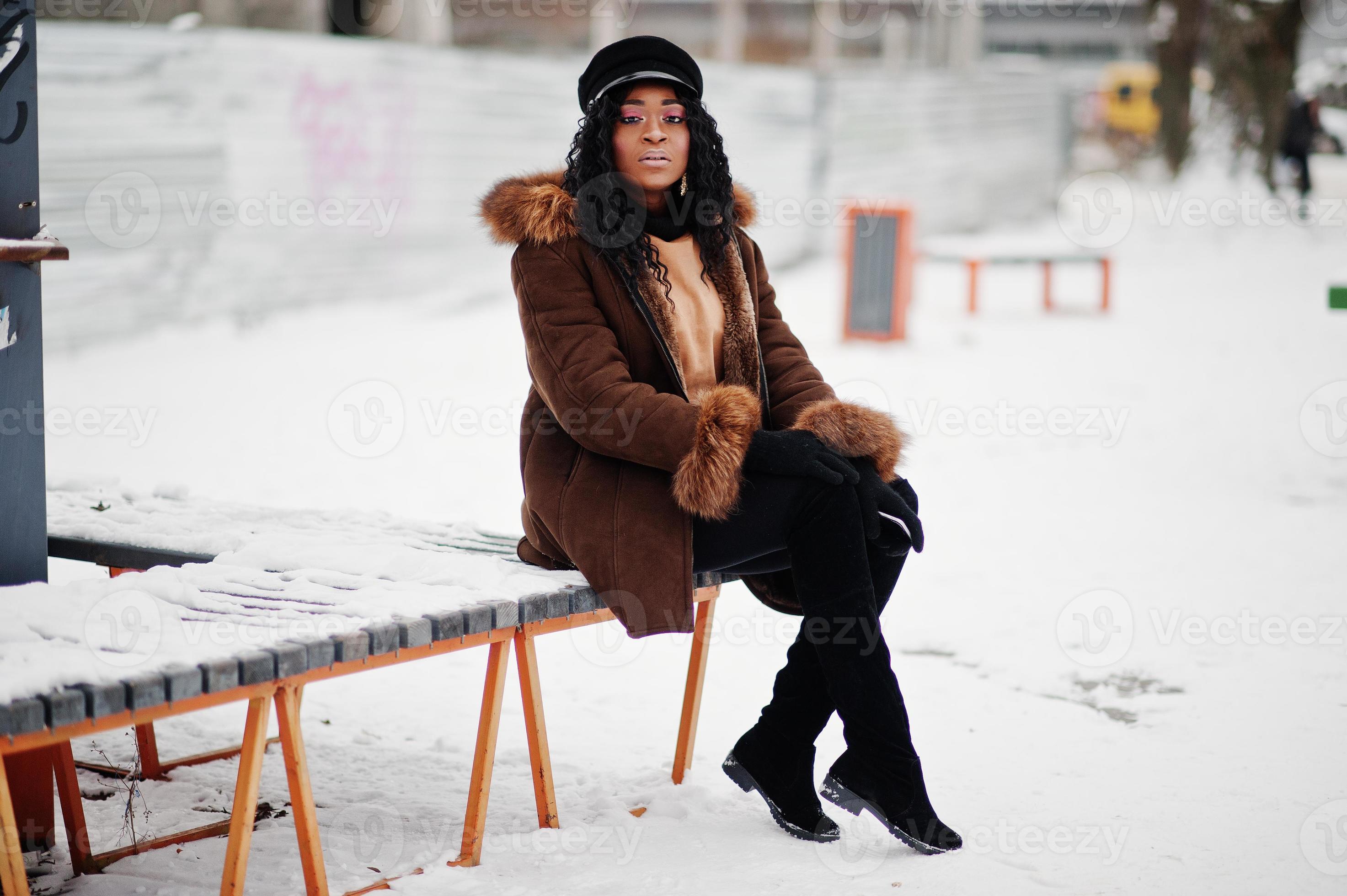 African american woman in sheepskin coat and cap posed at winter day  against snowy background, sitting on bench. 10417071 Stock Photo at Vecteezy