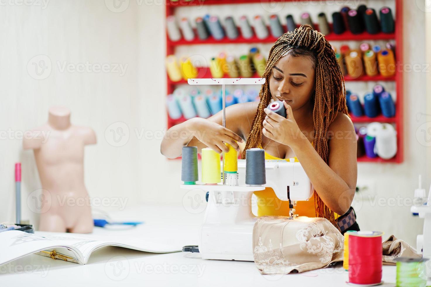 Young african dressmaker woman sews clothes on sewing machine and selects threads at tailor office. Black seamstress women. photo