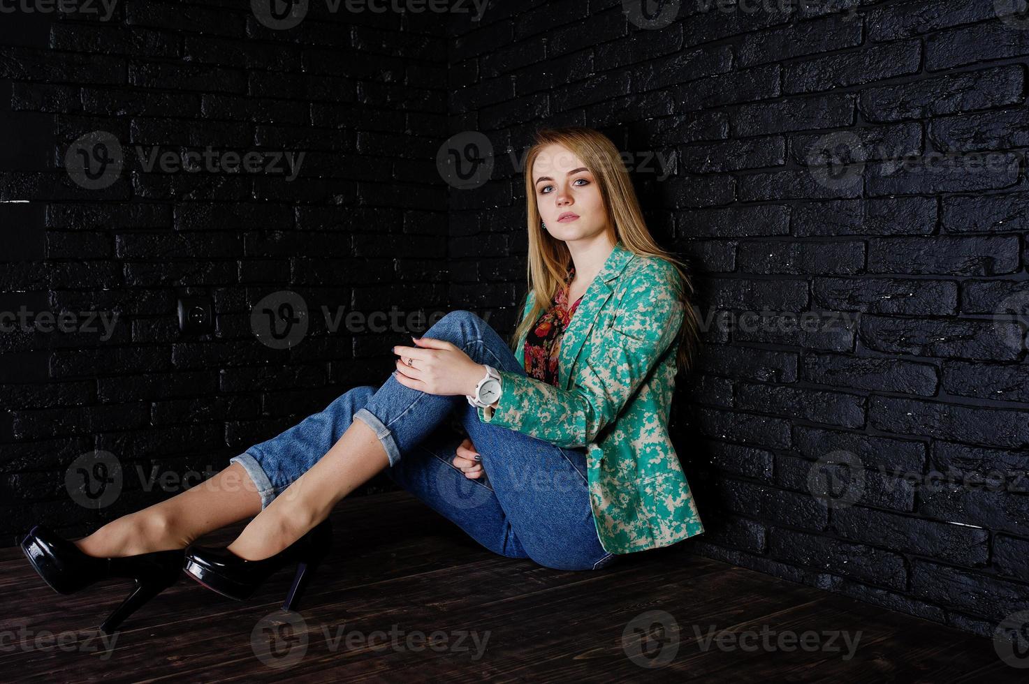 chica rubia con estilo en chaqueta y jeans contra la pared de ladrillo negro en el estudio. foto