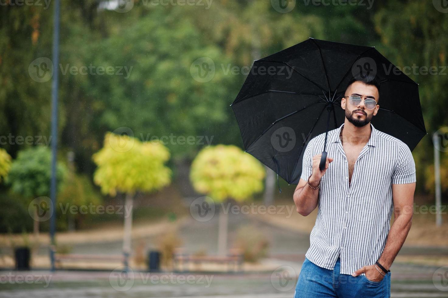 Fashionable tall arab beard man wear on shirt, jeans and sunglasses with umbrella posed at rain on park square. photo