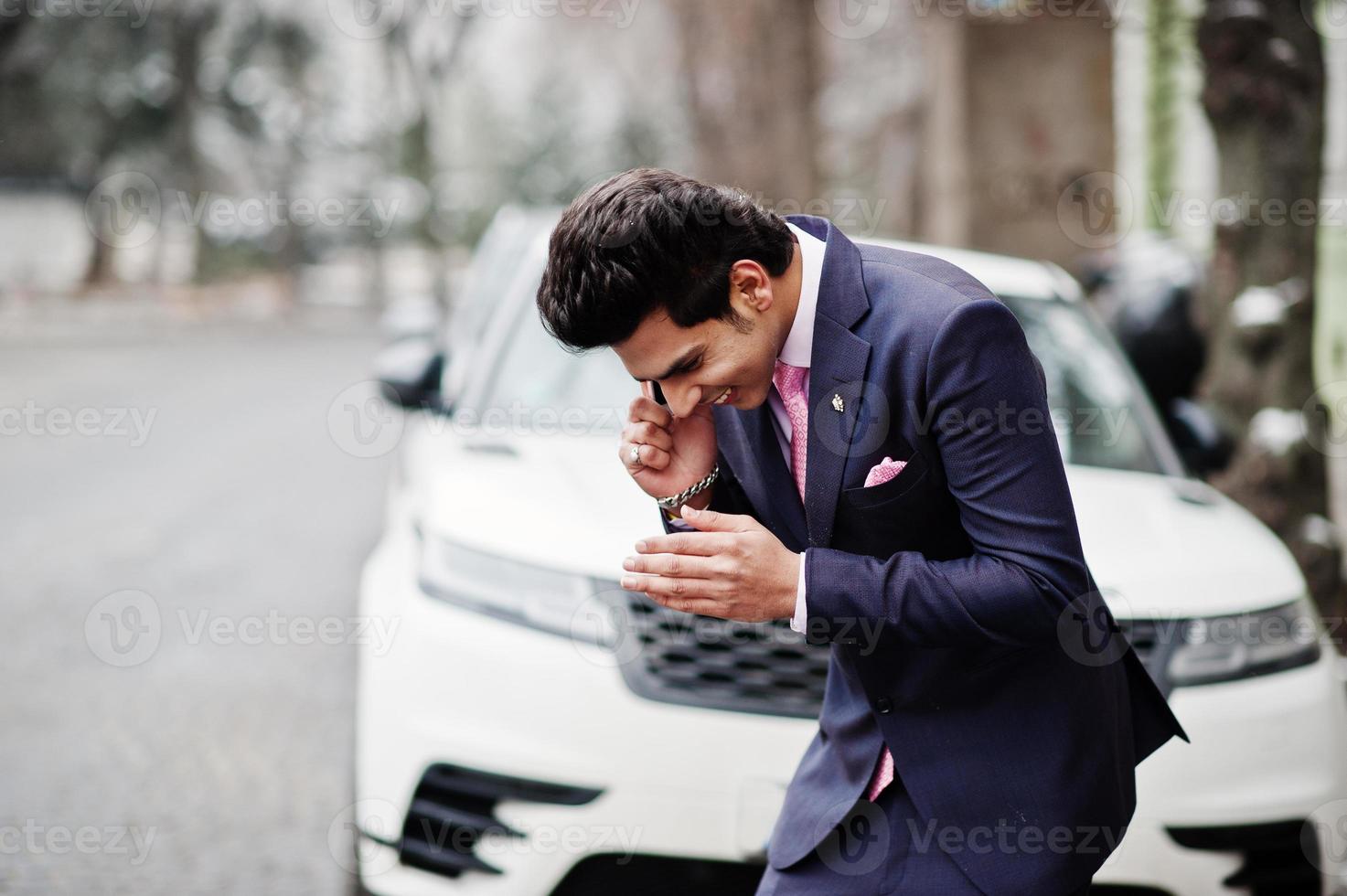 Elegant indian macho man model on suit and pink tie posed against white business car and speaking on phone. photo