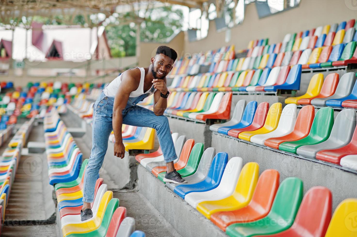 apuesto hombre afroamericano en overoles de jeans posados en sillas de colores en el estadio. retrato de hombre negro de moda. foto