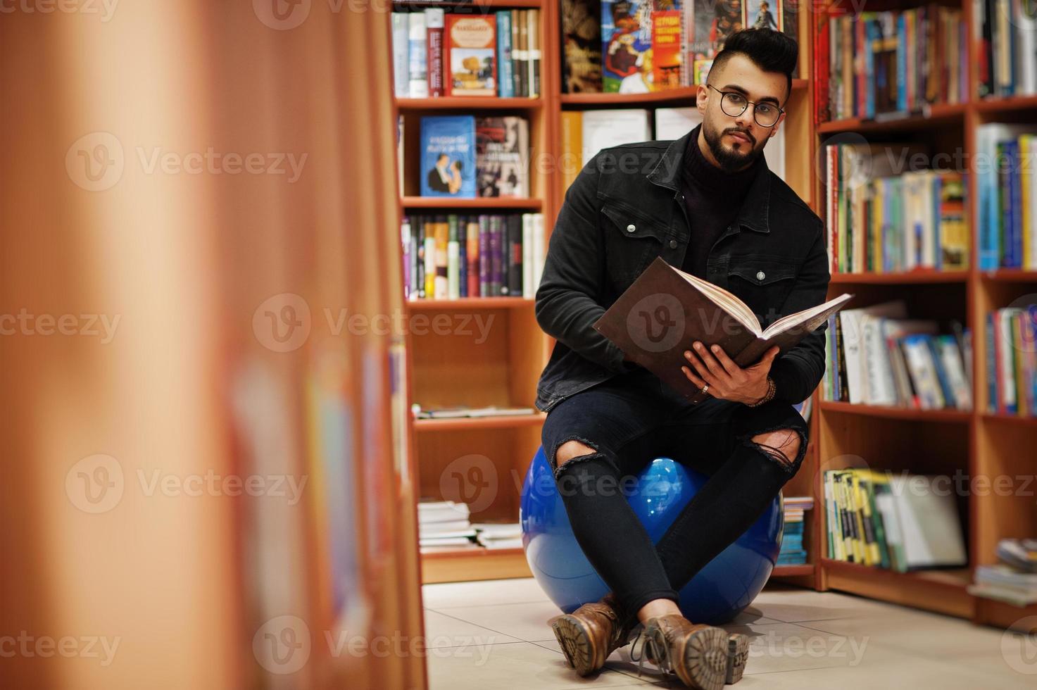 Tall smart arab student man, wear on black jeans jacket and eyeglasses, sitting at library and read book at hands. photo