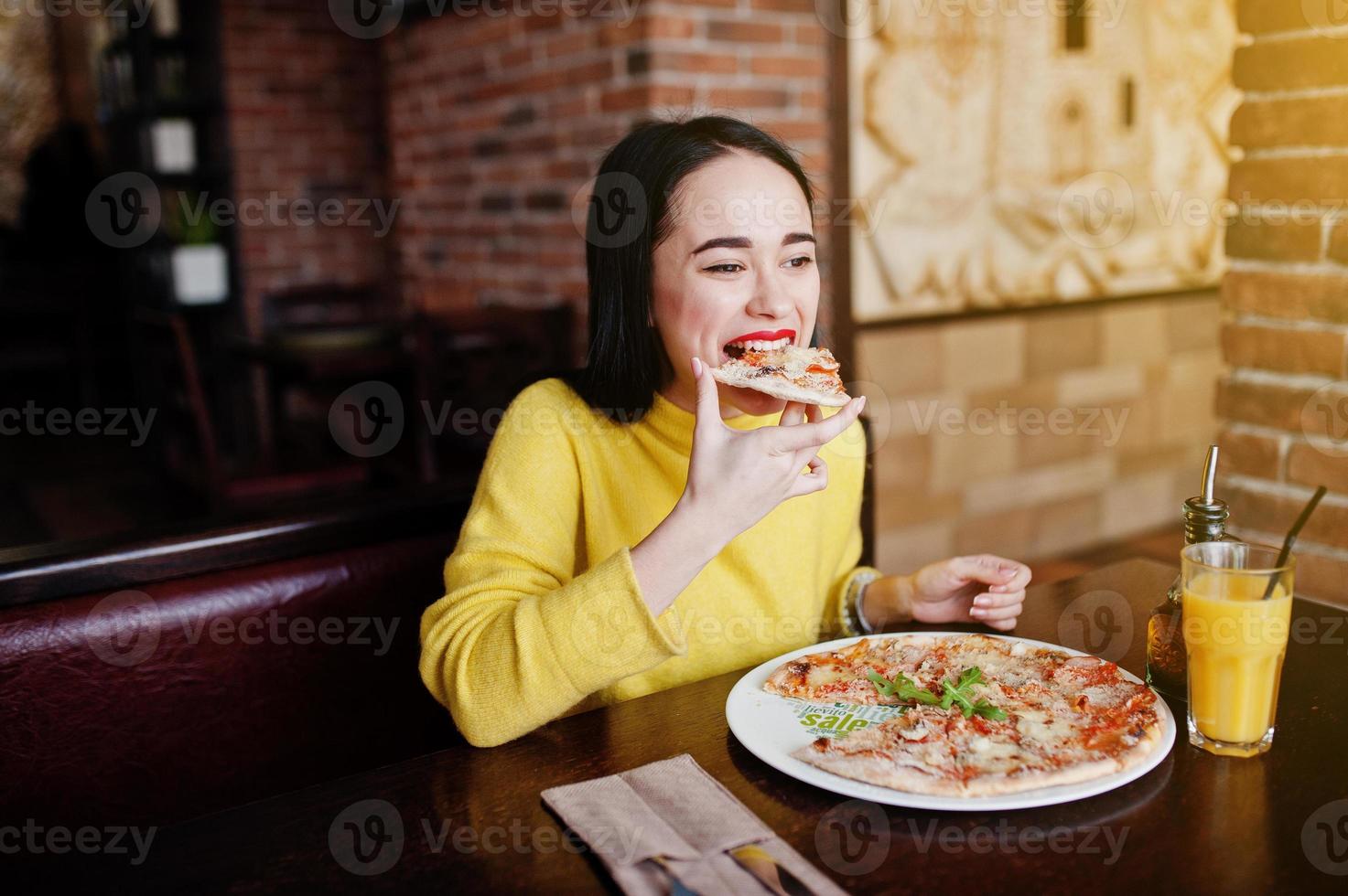 chica morena divertida en suéter amarillo comiendo pizza en el restaurante. foto