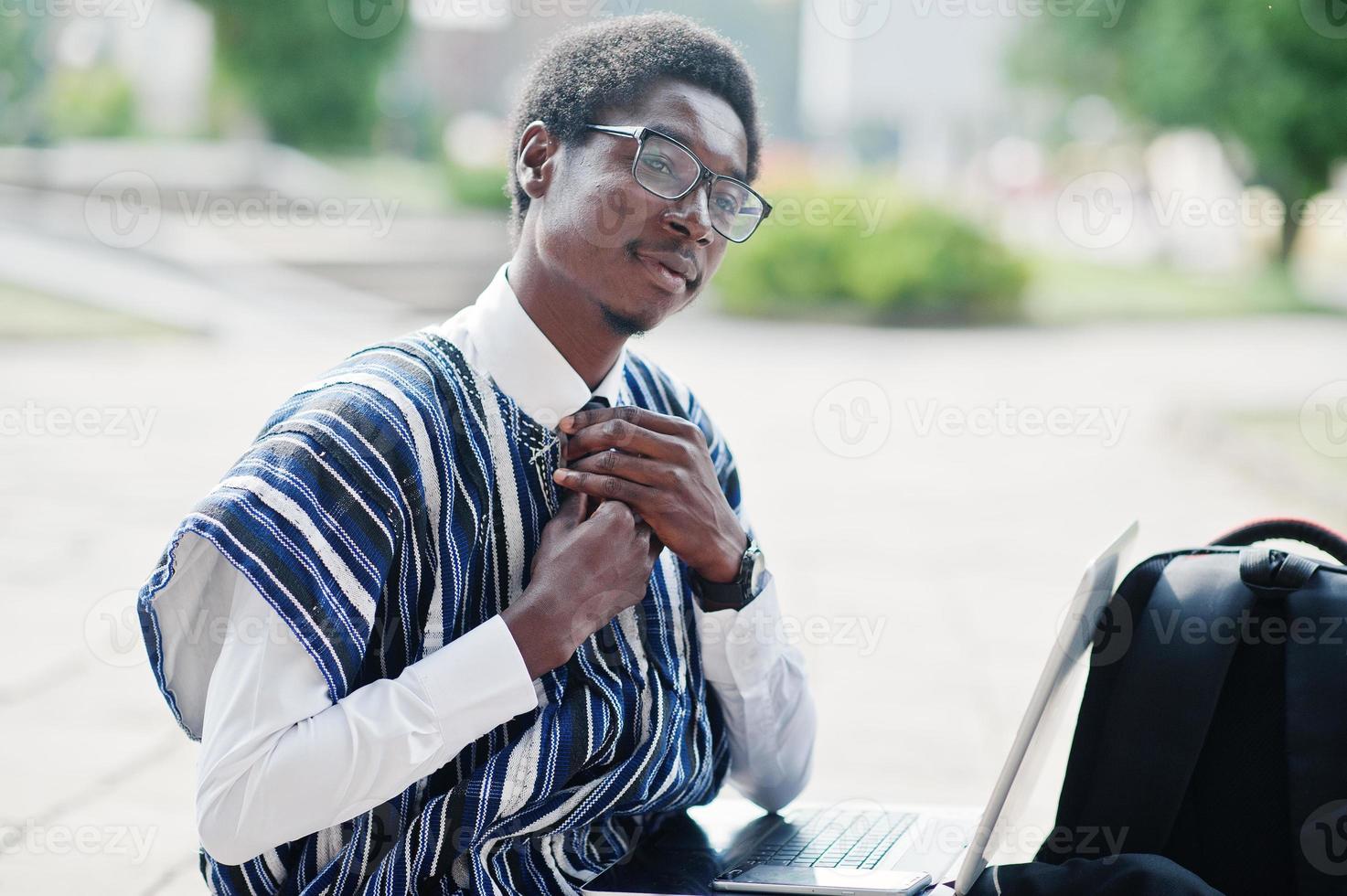 African man student in traditional clothes and glasses with backpack, working at laptop outdoor. photo