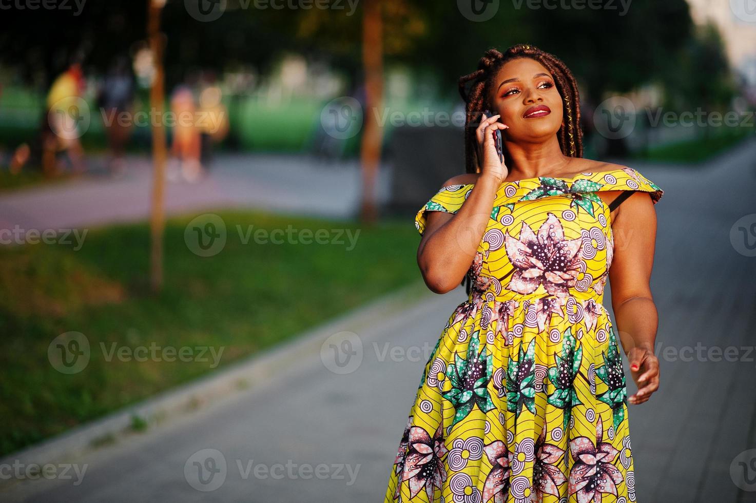 Cute small height african american girl with dreadlocks, wear at coloured yellow dress, posed at sunset, walking and speaking on cellphone. photo