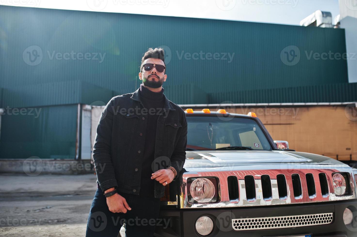 Fashion rich beard Arab man wear on black jeans jacket and sunglasses posed against big black suv car. Stylish, succesful and fashionable arabian model guy. photo