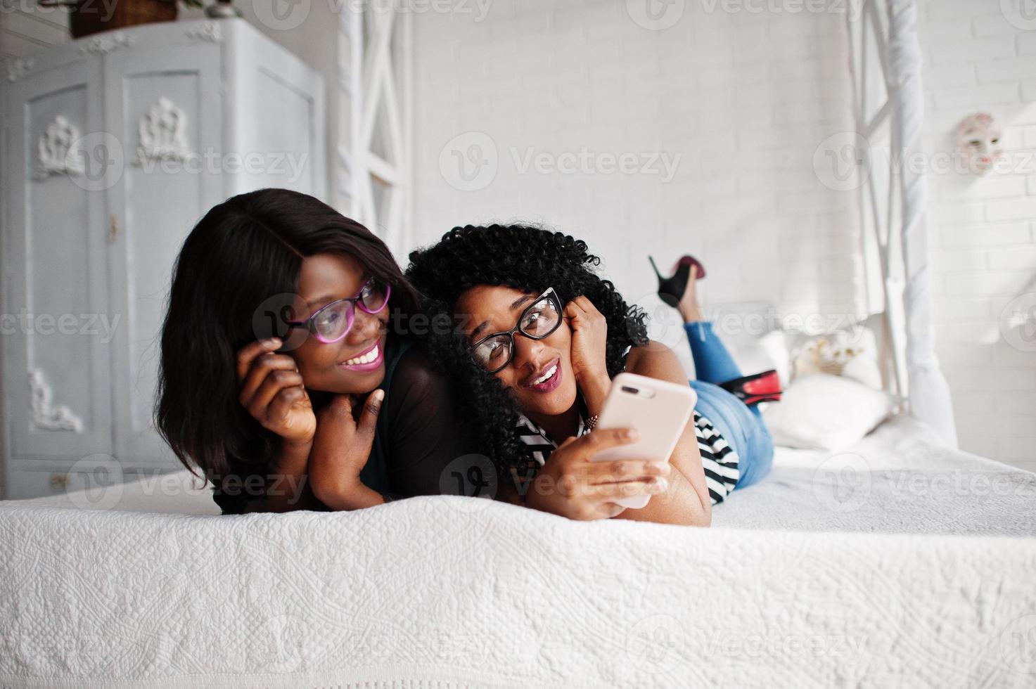 dos amigas africanas usan anteojos acostados en la habitación blanca interior de la cama y mirando el teléfono móvil. foto