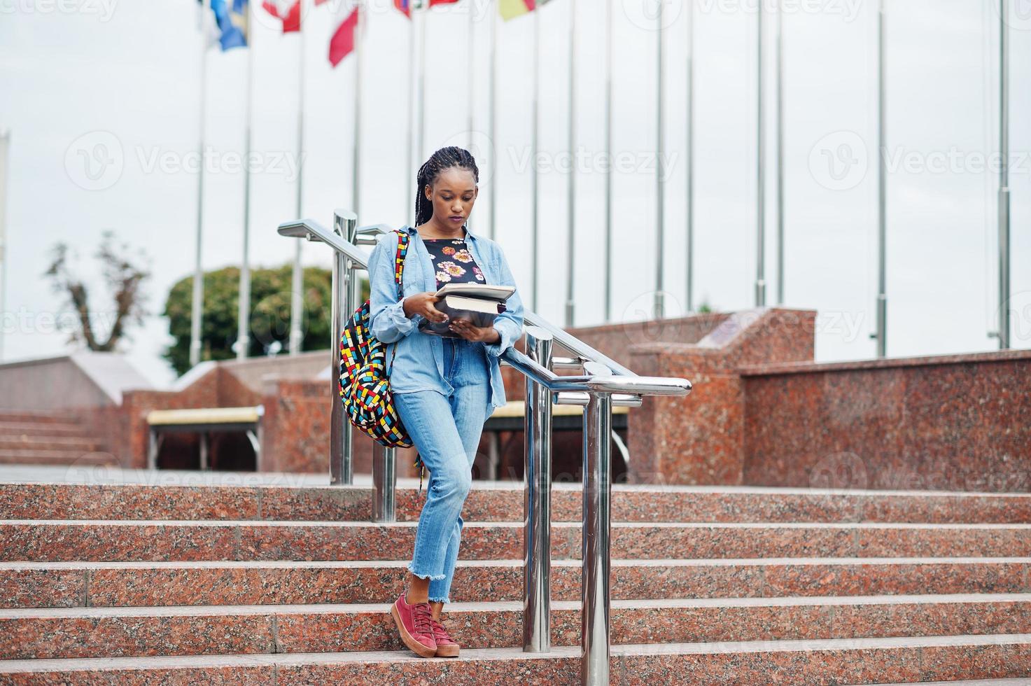 African student female posed with backpack and school items on yard of university, against flags of different countries. photo