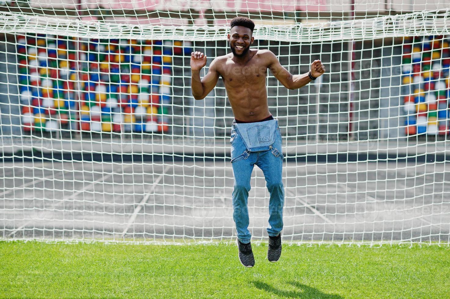 Handsome sexy african american muscular bare torso man at jeans overalls posed at green grass agasinst football gates of stadium field. Fashionable black man portrait. photo