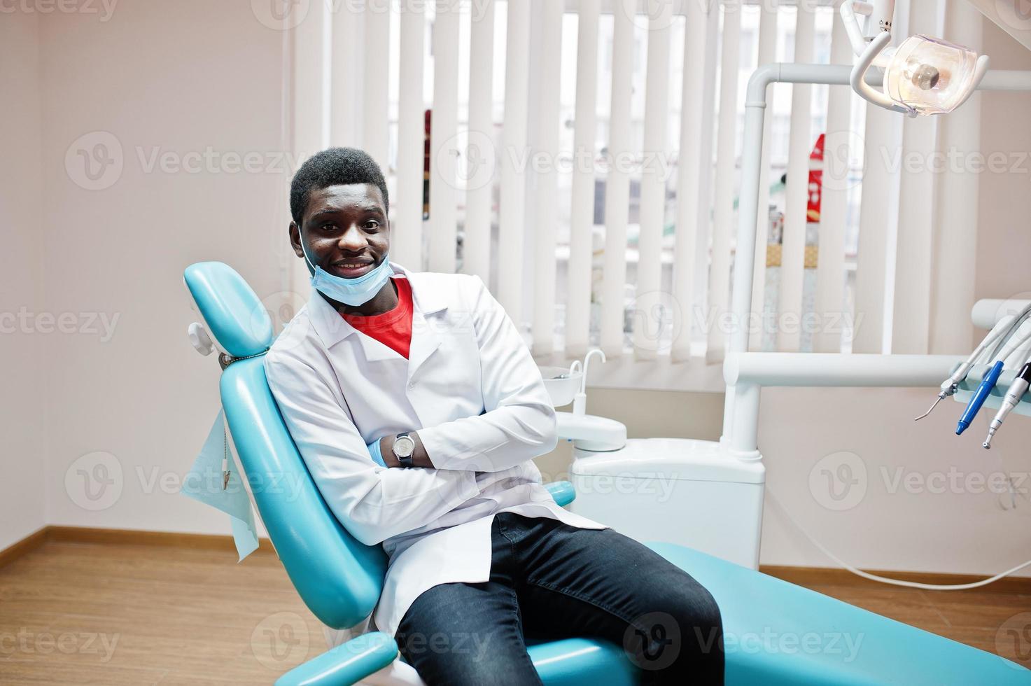 médico afroamericano enmascarado con brazos cruzados sentado en la silla del dentista en la clínica dental. foto