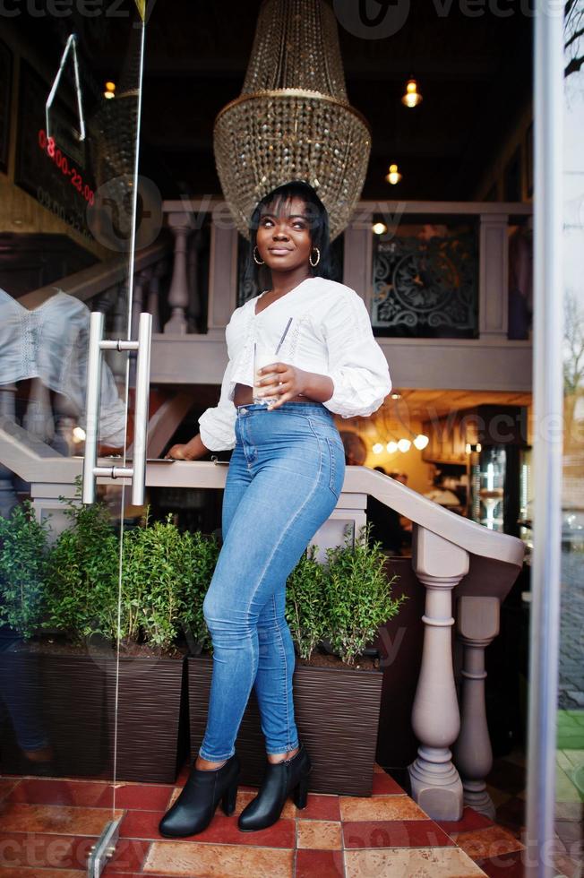 Stylish african american women in white blouse and blue jeans posed at cafe with caramel latte. photo