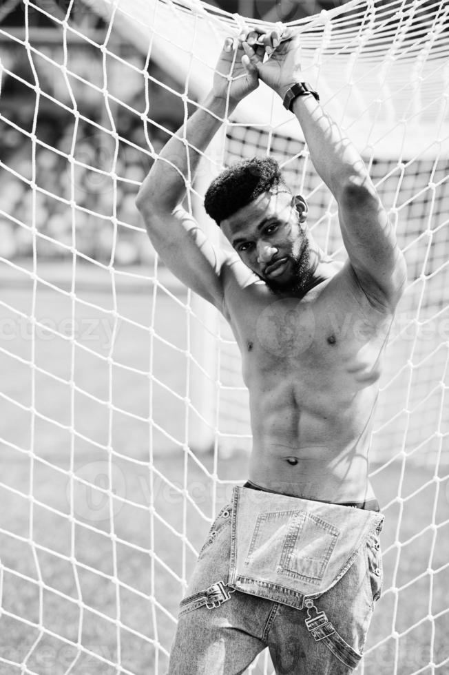 Handsome sexy african american muscular bare torso man at jeans overalls posed at green grass agasinst football gates of stadium field. Fashionable black man portrait. photo