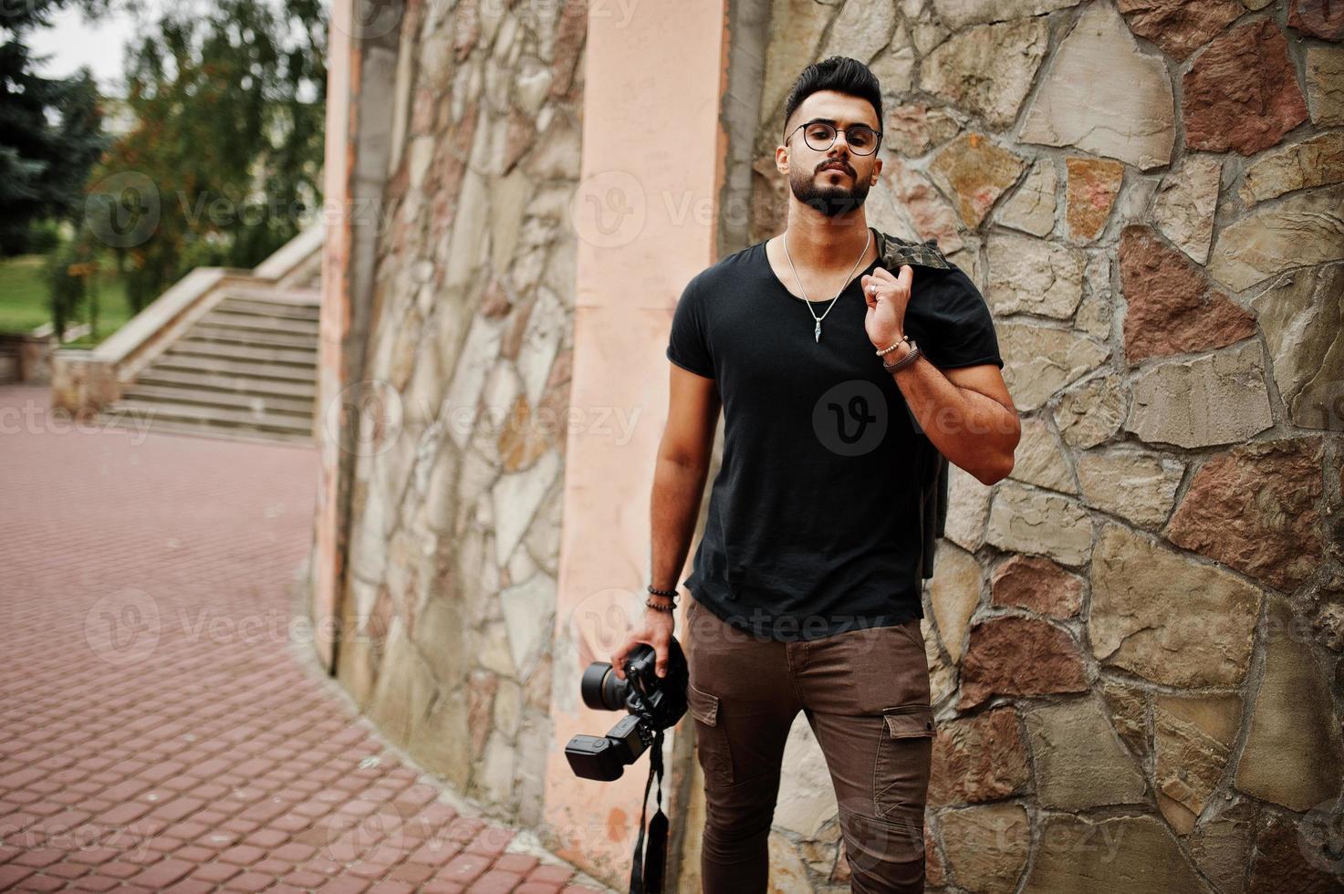 Impresionante y hermoso fotógrafo de hombre macho con barba árabe alta con gafas y camiseta negra con cámara profesional en las manos. foto