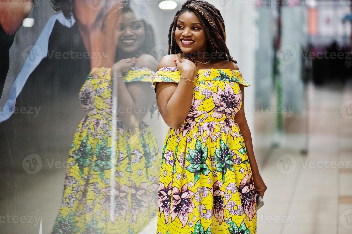 linda chica afroamericana de pequeña altura con rastas, vestida con un vestido amarillo de color, posada en el escaparate de la tienda en el centro comercial. foto