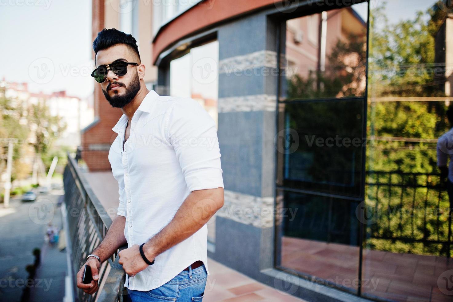 Stylish tall arabian man model in white shirt, jeans and sunglasses posed at street of city. Beard attractive arab guy against modern building. photo