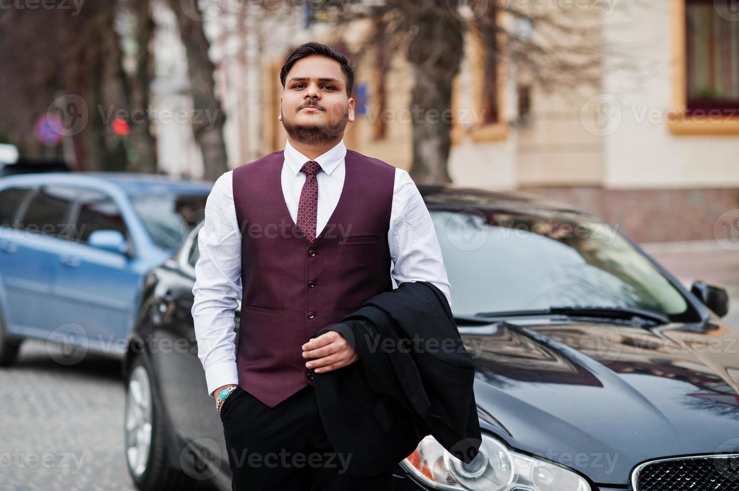 Stylish indian businessman in formal wear vest suit standing against black business car on street of city. photo