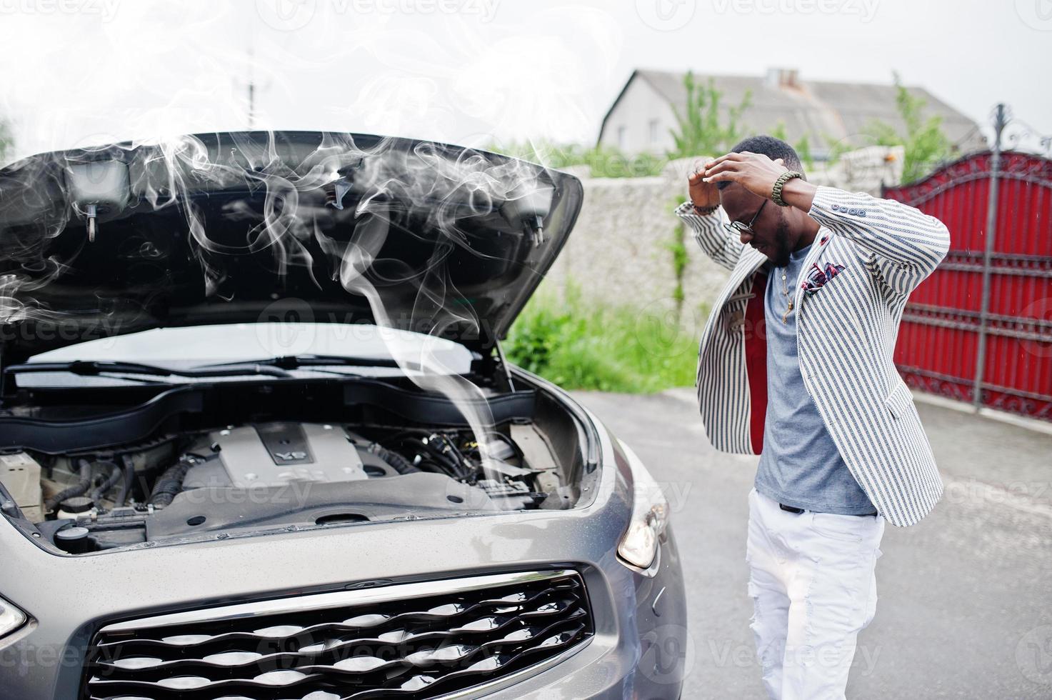 Un hombre afroamericano elegante y rico parado frente a un auto todoterreno roto necesita ayuda para mirar debajo del capó abierto con humo. foto