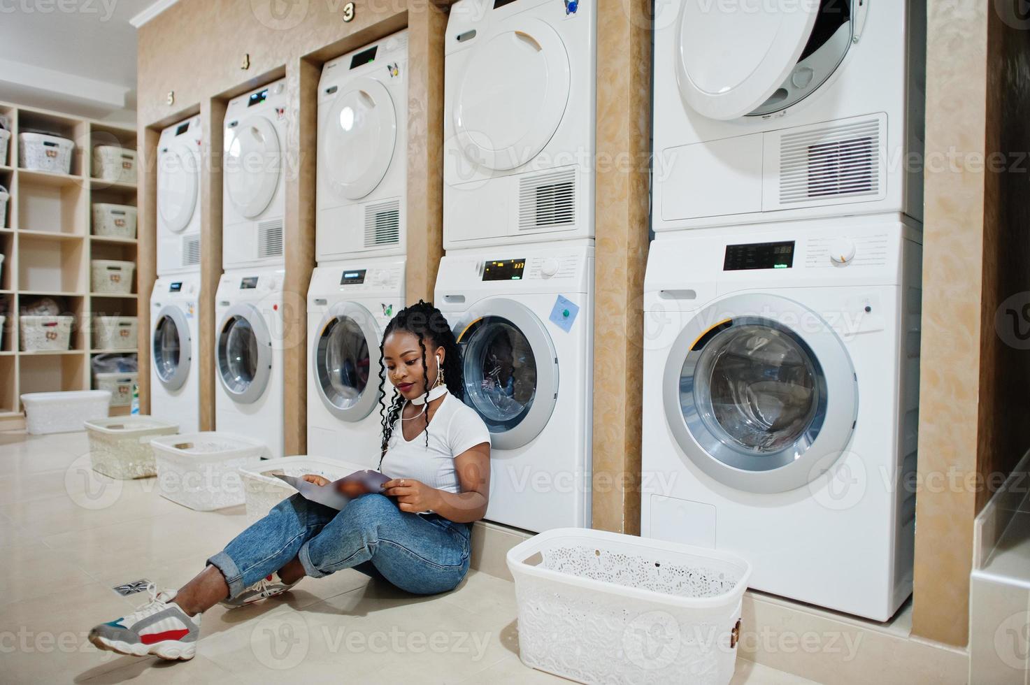 Cheerful african american woman sitting with earphones and read magazine near washing machine in the self-service laundry. photo