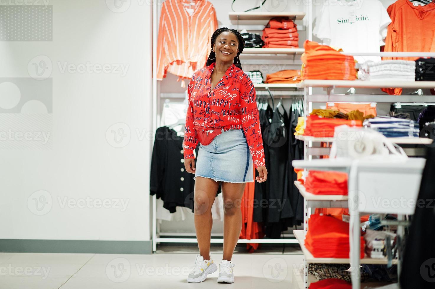 Modish african american woman in red shirt, jeans skirt with crossbody bag posed at clothes store. It's time for shopping. photo