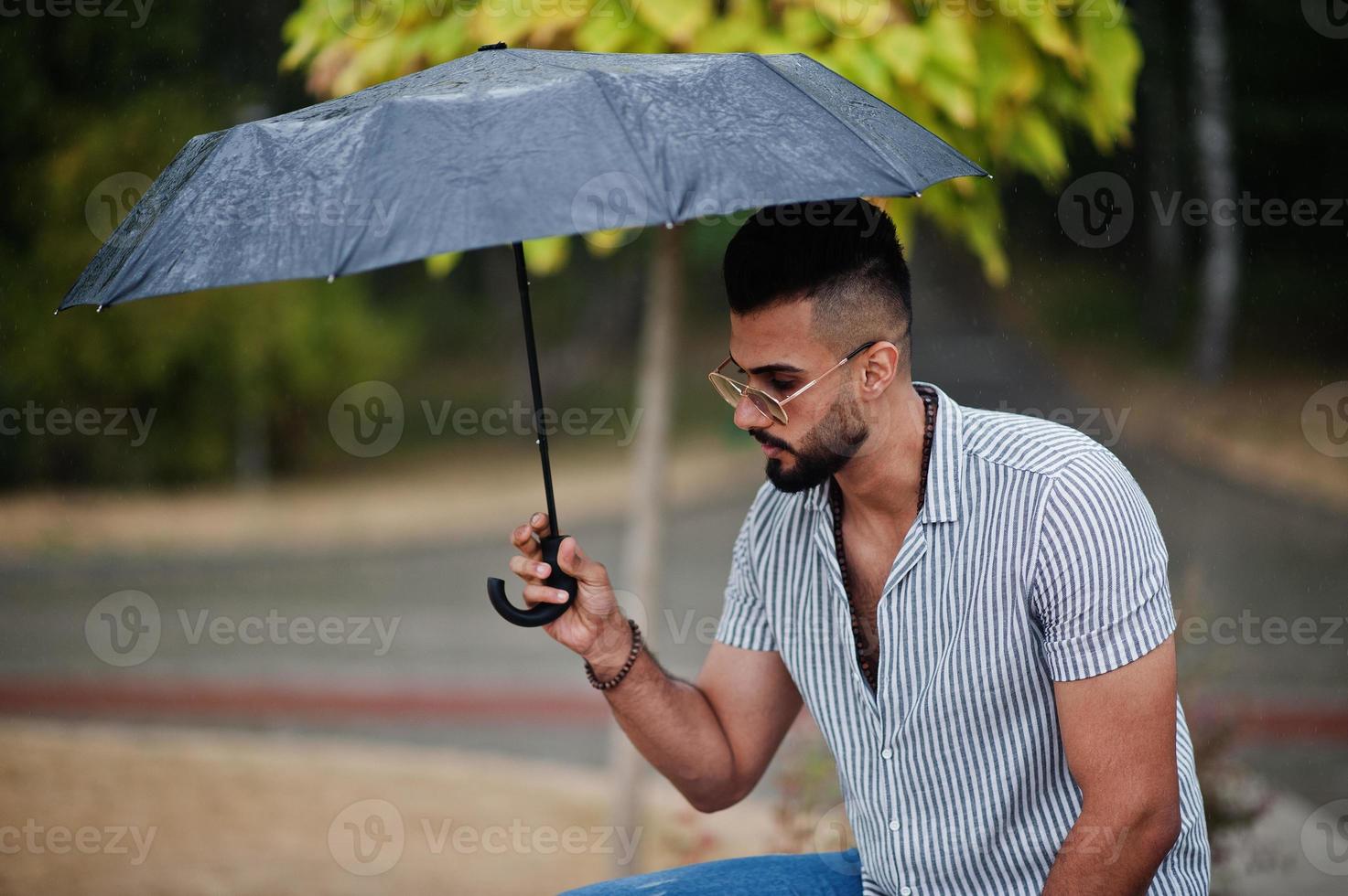 Fashionable tall arab beard man wear on shirt, jeans and sunglasses with umbrella posed at rain on park square. photo